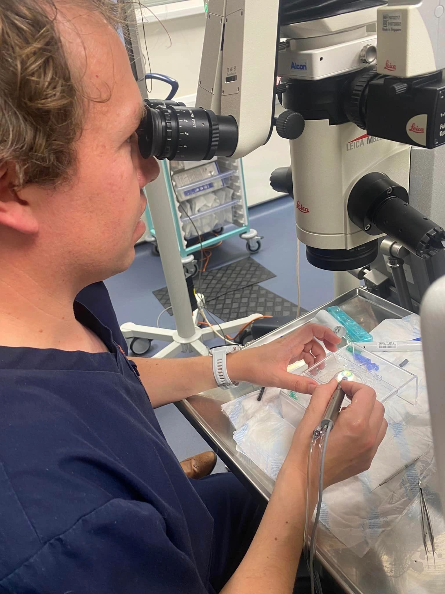 A Fulgoni Opticians team member in their navy Fulgoni-branded scrubs at the Leica M822 surgical microscope, practising intraocular lens technique at The Wye Clinic Hereford in 2023